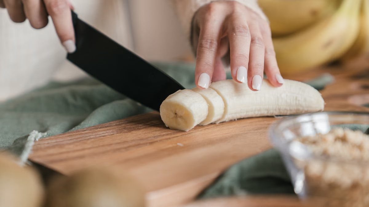 A close-up view of a hand slicing a banana on a wooden chopping board.